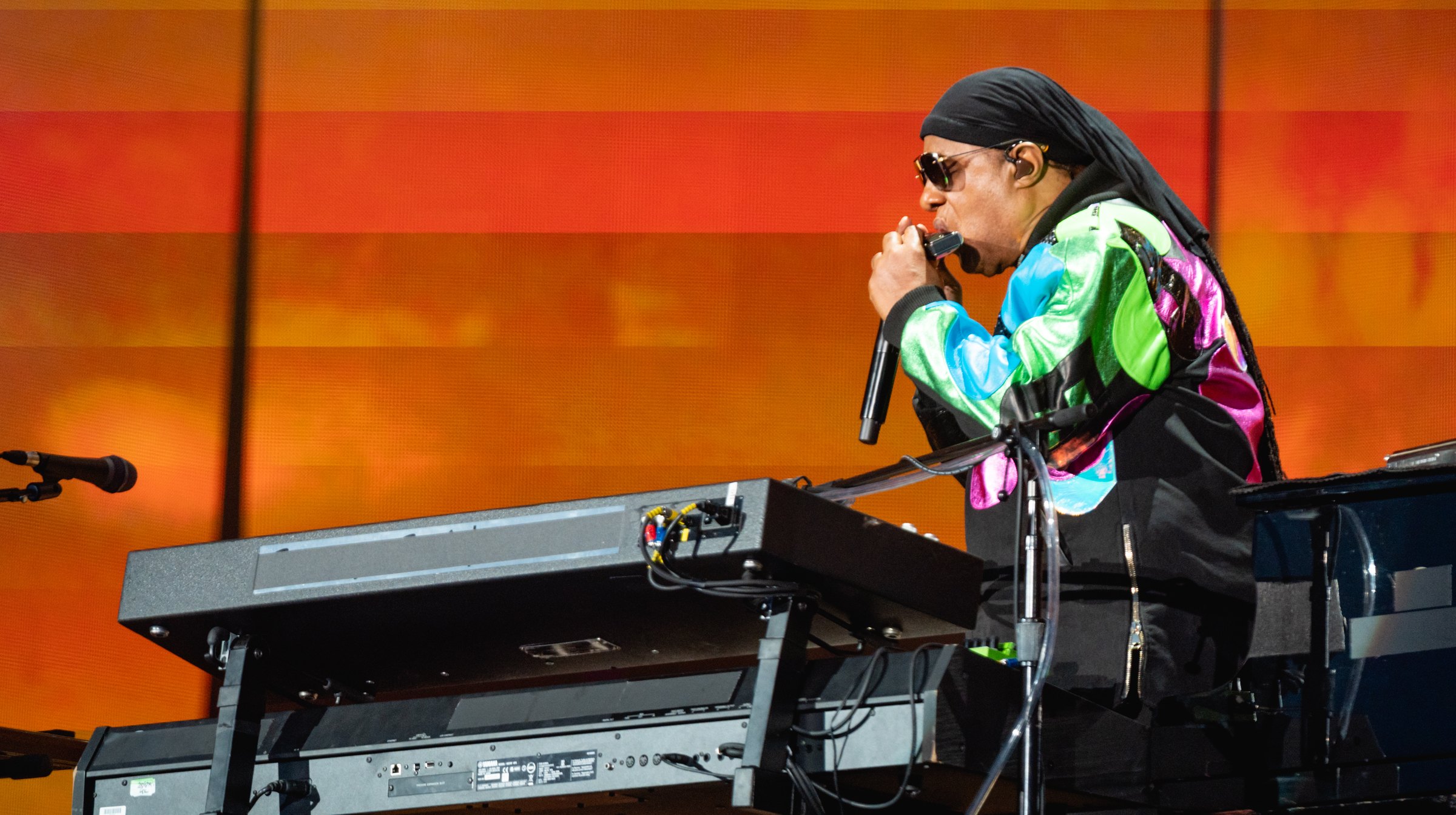 Stevie Wonder playing harmonica behind his keyboards in front of a glowing orange stage screen